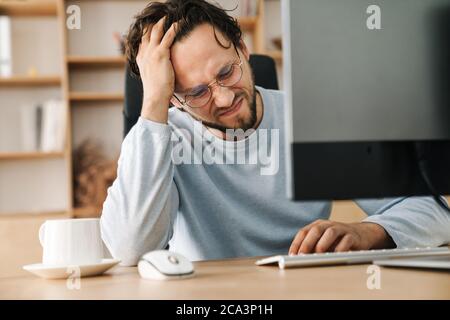 Image of unhappy bearded programmer man wearing eyeglasses working with computer in office Stock Photo