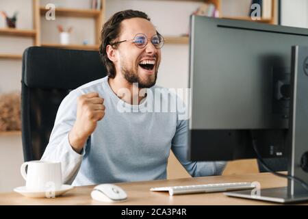 Image of laughing programmer man making winner gesture while working with computer in office Stock Photo