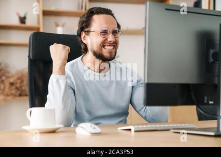 Image of laughing programmer man making winner gesture while working with computer in office Stock Photo
