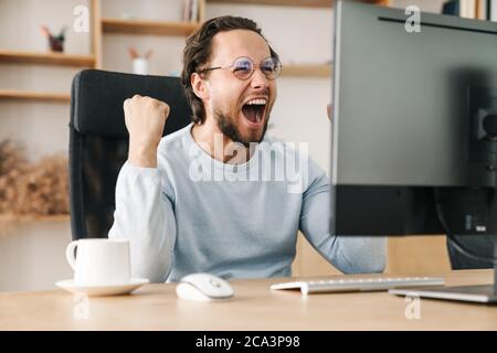 Image of laughing programmer man making winner gesture while working with computer in office Stock Photo
