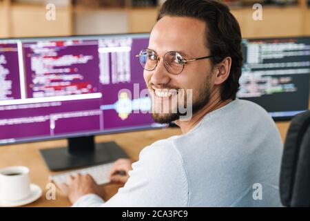 Image of smiling unshaven programmer man wearing eyeglasses working with computers in office Stock Photo