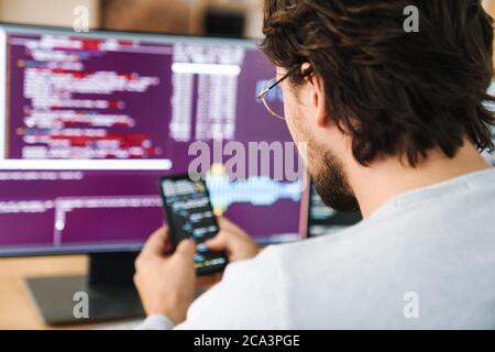 Image of unshaven focused programmer man using smartphone while working with computers in office Stock Photo
