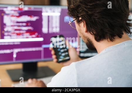 Image of unshaven focused programmer man using smartphone while working with computers in office Stock Photo