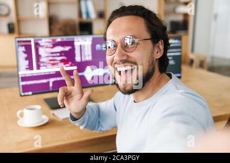 Image of unshaven excited programmer man gesturing peace sign while taking selfie photo in office Stock Photo