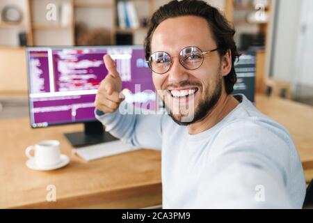 Image of unshaven excited programmer man pointing finger at camera while taking selfie photo in office Stock Photo
