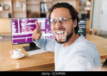 Image of unshaven excited programmer man pointing finger at camera while taking selfie photo in office Stock Photo