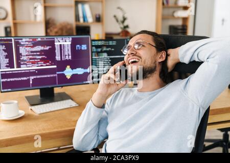 Image of handsome joyful programmer man laughing and talking on cellphone while working in office Stock Photo