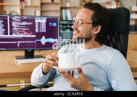 Image of handsome joyful programmer man smiling and drinking coffee while working in office Stock Photo