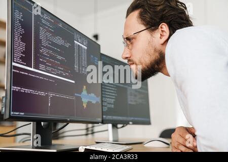 Image of concentrated unshaven programmer man wearing eyeglasses working with computers in office Stock Photo