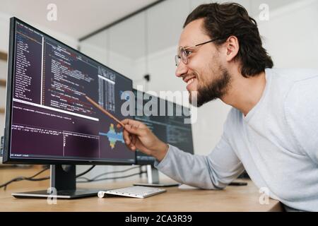 Image of smiling unshaven programmer man wearing eyeglasses working with computers in office Stock Photo