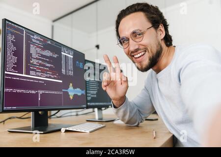Image of smiling unshaven programmer man showing peace sign while working with computers in office Stock Photo