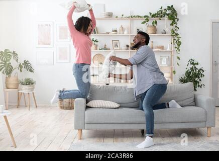 Young Spouses Leisure. Playful Black Couple Having Pillowfight In Living Room Stock Photo