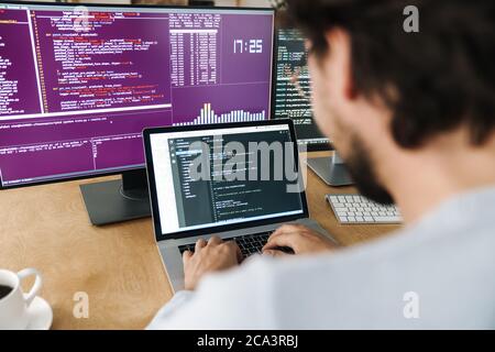 Image of focused caucasian programmer man wearing eyeglasses working with computers in office Stock Photo