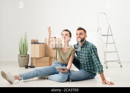 Positive young couple sitting on floor in new apartment with moving stuff and using design app on tablet while discussing interior Stock Photo