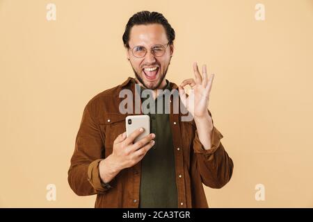 Image of excited bristle man in eyeglasses gesturing ok sign and using cellphone isolated over beige background Stock Photo