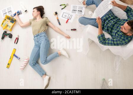 Above view of exhausted young woman with paint roller lying among repair tools while her husband analyzing house plan Stock Photo