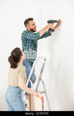 Excited young bearded man standing on ladder and turning screw into wall while hanging picture on wall with wife Stock Photo