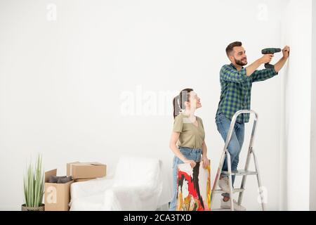 Positive young man standing on step ladder and using electric screwdriver while inserting screw for picture Stock Photo