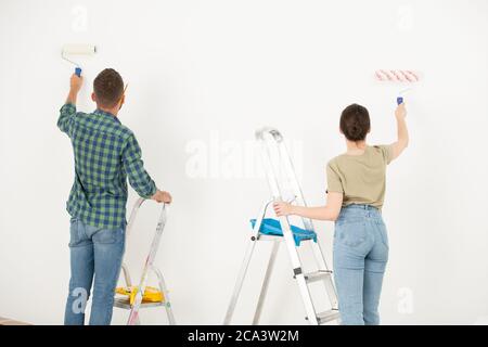 Rear view of young couple painting wall white using paint roller while remodeling their old room Stock Photo