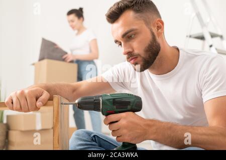 Concentrated young bearded man assembling furniture using cordless screwdriver in new apartment Stock Photo