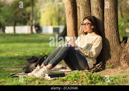 Female student using phone, sitting under tree in park Stock Photo