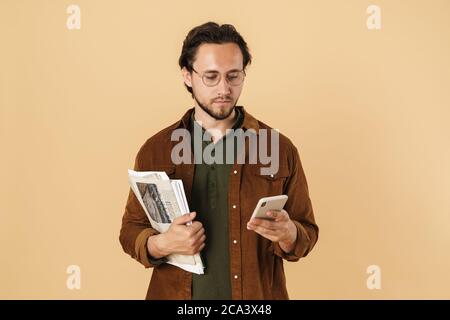 Image of serious unshaven man using mobile phone while holding newspapers isolated over beige background Stock Photo