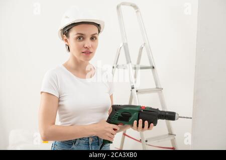 Portrait of content young woman in hardhat using electric drill while doing renovation in new flat Stock Photo