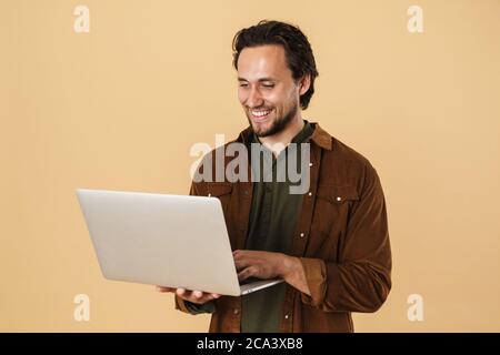 Image of cheerful bearded man smiling and typing on notebook isolated over beige background Stock Photo