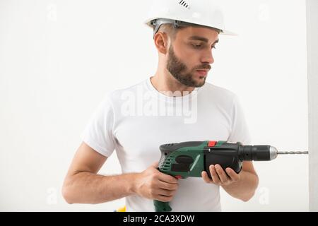 Concentrated young bearded man in construction hardhat drilling hole into wall while making renovation in new flat Stock Photo