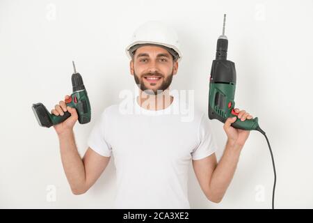 Portrait of smiling young bearded repairman in hardhat holding cordless screwdriver and drill against white wall Stock Photo