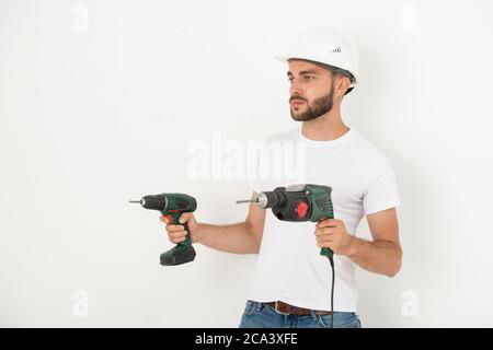 Serious young bearded repairman in hardhat holding power hand tools while preparing for remodeling in flat Stock Photo