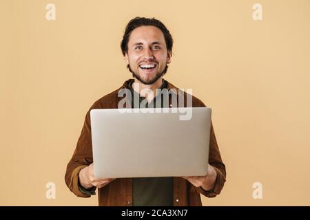Image of pleased unshaven man smiling while using laptop isolated over beige background Stock Photo