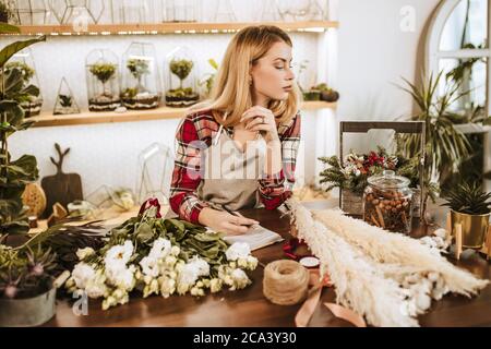 young caucasian lady florist make notes at work, wearing apron on red checkered casual shirt, lady surrounded by green plants. use notebook Stock Photo