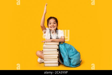 School Girl Raising Hand Sitting At Book Stack, Studio Shot Stock Photo