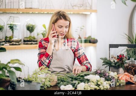 attractive caucasian woman florist in red checkered shirt with smartphone, talk with friend while no customers in flowers shop or store Stock Photo