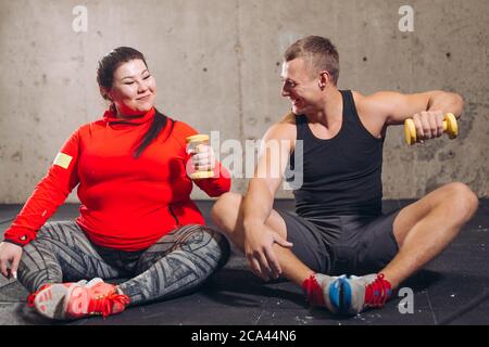 positive overweight woman and fit guy doing exercises while sitting on the floor. full length photo. funny learning. studing. Stock Photo