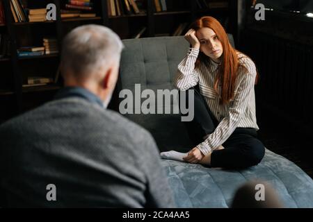 Frustrated young red-haired lady in casual clothing is having therapy session with psychologist. Stock Photo