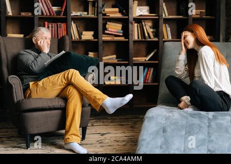 Happy red-haired young woman patient is telling about her mental health lying in comfortable sofa. Stock Photo