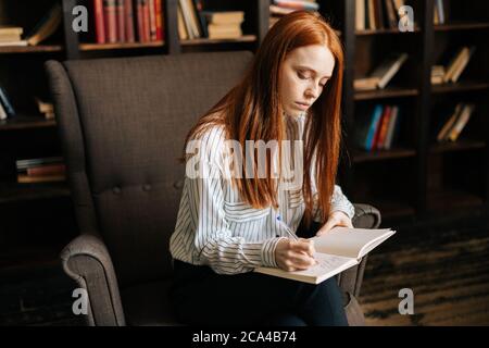 Focused young businesswoman with red hair making notes in notebook at home office. Stock Photo