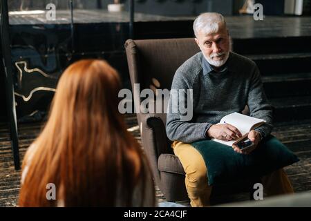 Over the shoulder shot of red-haired young woman patient speaking with mature man psychologist.  Stock Photo