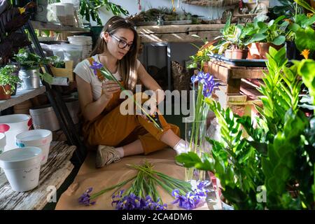 Small business. Florist woman surrounded by tropical plants cutting the stem of irises flowers using secateurs, sitting on the floor in flower store. Stock Photo