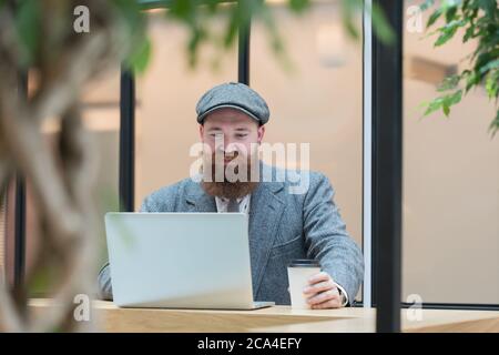 Portrait of brutal bearded hipster man wearing wool blazer, cap working on laptop sitting in cafe/ restaurant, drinking coffee from a paper cup, indoo Stock Photo