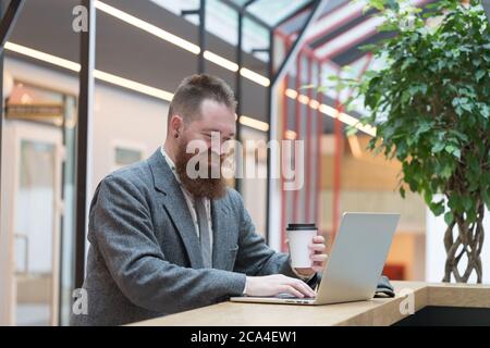 Portrait of brutal bearded hipster man wearing wool blazer working on laptop sitting in cafe/ restaurant, drinking coffee from a paper cup, indoor. Di Stock Photo