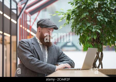 Portrait of brutal bearded hipster man wearing wool blazer, cap working on laptop sitting in cafe/ restaurant, indoor. Distance job, remote work Stock Photo