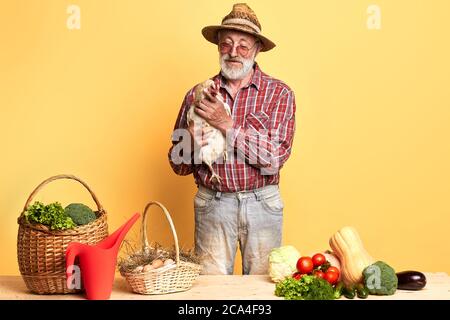 Senior male farmer came to market with fresh vegetables and eggs from his local farm, holding with care his dunghill hen, waiting for buyers, smiling Stock Photo