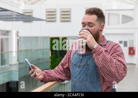 Brutal bearded self employed worker in blue overalls, checked shirt, drinking coffee from a paper cup, using smartphone. Time to take break. Hipster b Stock Photo