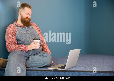 Portrait of brutal bearded hipster man in blue overalls, checked shirt, drinking coffee from a paper cup working on laptop sitting near the wall. Dist Stock Photo