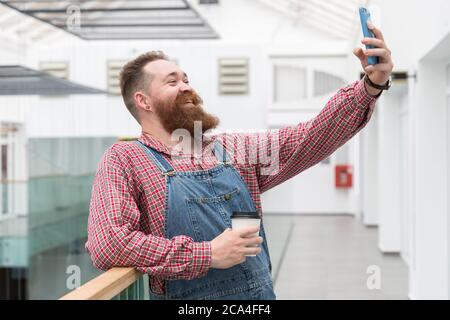 Smiling bearded barber worker in blue overalls, checked shirt, drinking coffee from a paper cup, talking on a video call or taking a selfie on smartph Stock Photo