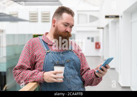Brutal bearded self employed worker in blue overalls, checked shirt, drinking coffee from a paper cup, using smartphone. Time to take break. Hipster b Stock Photo
