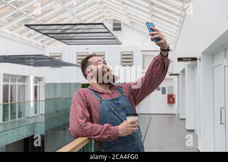 Smiling bearded barber worker in blue overalls, checked shirt, drinking coffee from a paper cup, talking on a video call or taking a selfie on smartph Stock Photo